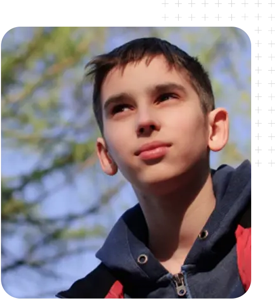 A young boy stands outdoors, wearing a navy hoodie with a red accent, surrounded by green trees and bright blue skies.